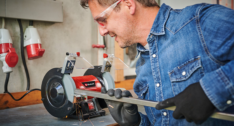 A man grinding a metal rod with an Einhell bench grinder in a workshop.